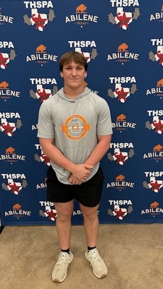 A man stands in front of a blue backdrop with text and logos, wearing a gray hoodie and black shorts.