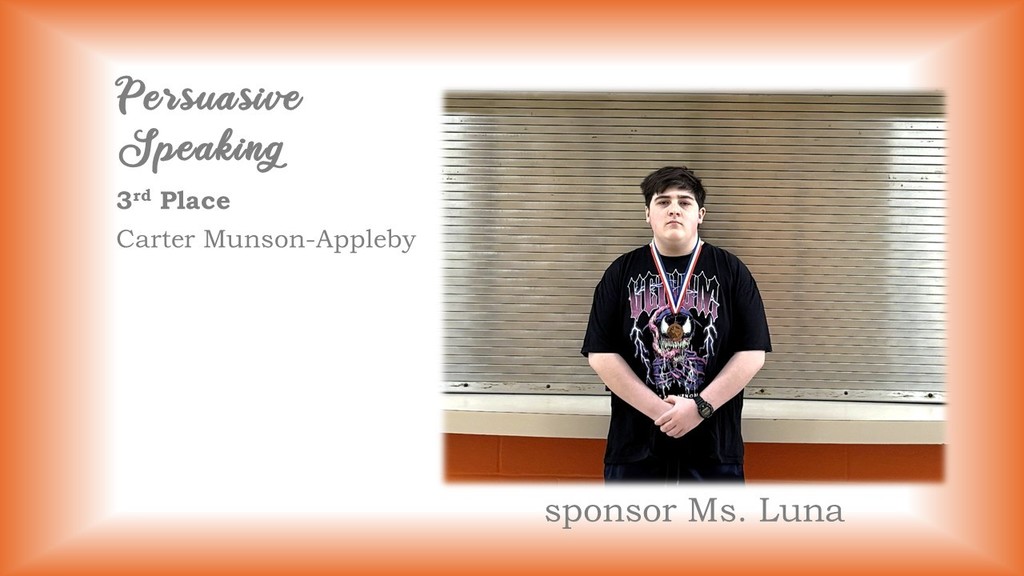 A person stands before a window with blinds, wearing a black t-shirt with a graphic design.