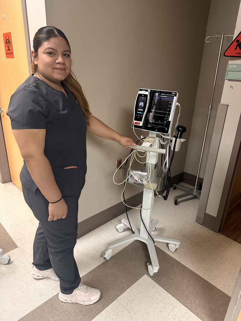 A student stands next to a medical monitor on a rolling cart in a hospital room.