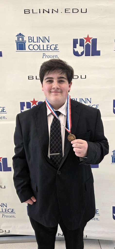A young man in a suit poses with a medal in front of a Blinn College banner.