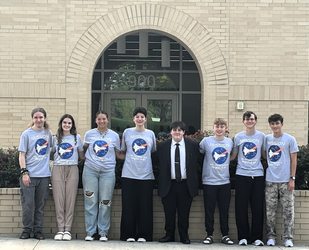 Seven people in matching shirts stand in front of a brick building, each wearing sneakers and some have ripped jeans. One student is in a black suit.