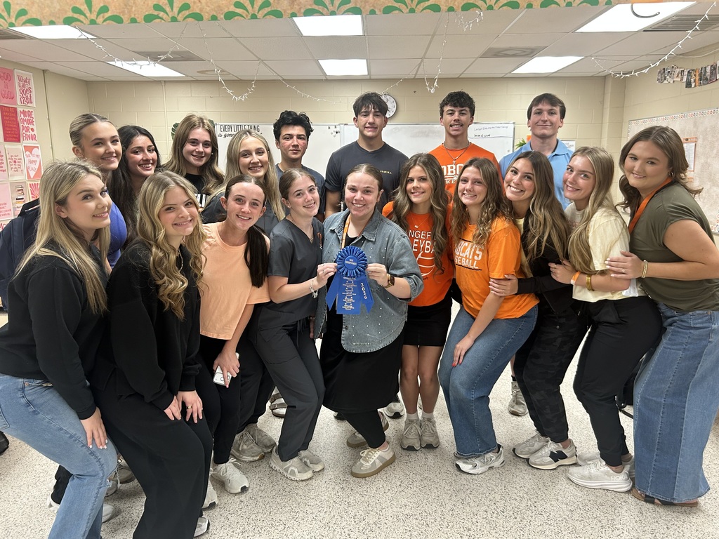 Group of people posing for a photo in a room. Holding a blue ribbon. Background includes a white floor and bulletin board.
