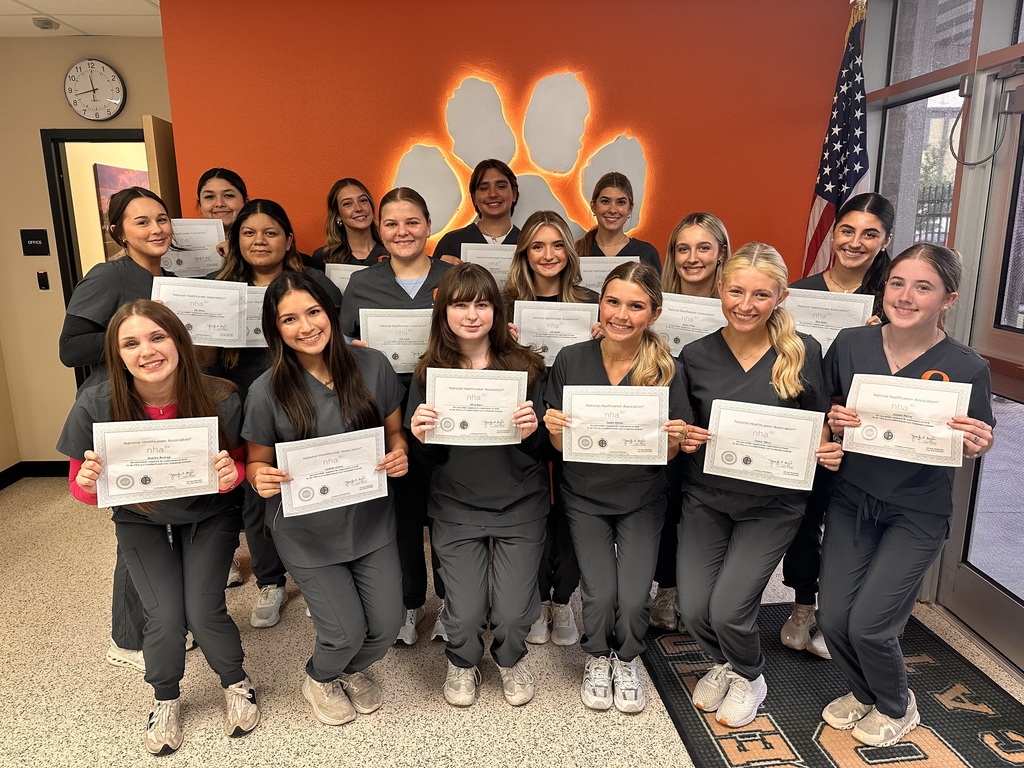 A group of students in matching uniforms hold certificates and pose for a photo in front of an orange wall with a paw.