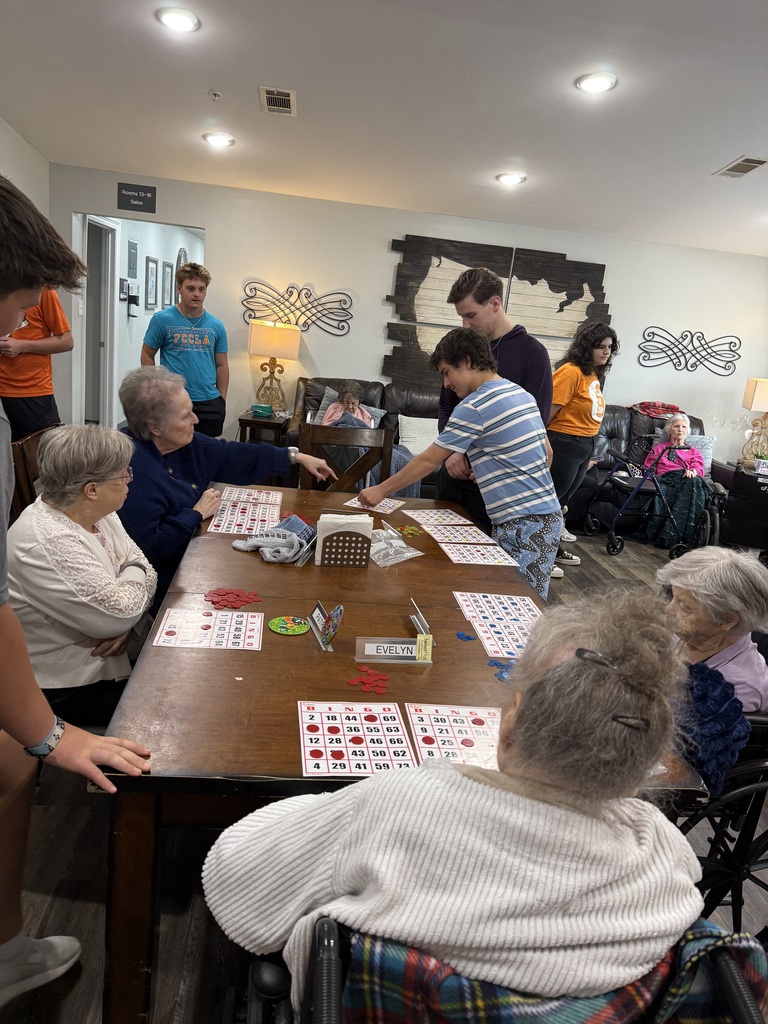 Several older people and younger adults engage in a table game. Two young men in blue shirts assist.