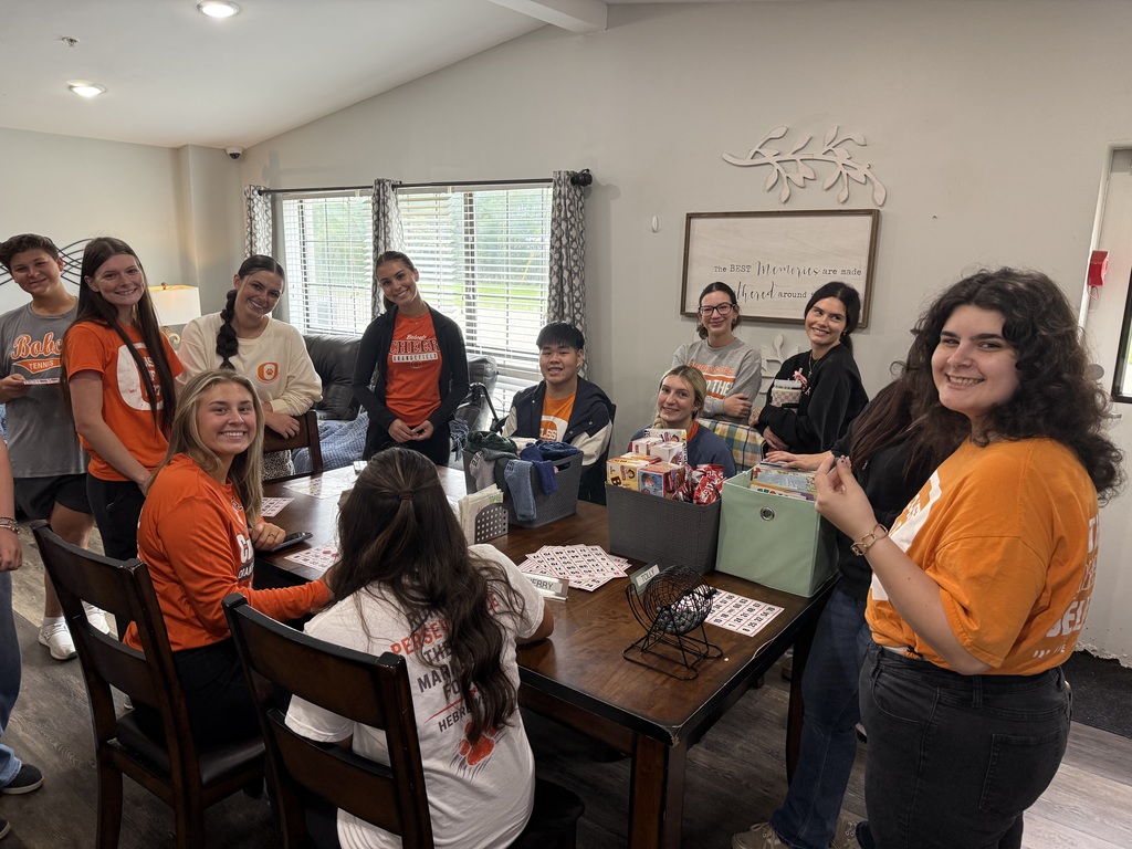 A group of people in orange and white shirts gather around a table with various items on it.