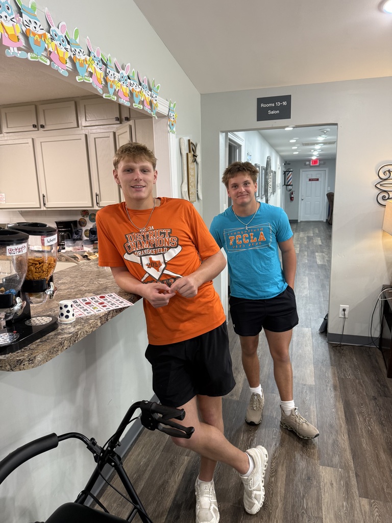 Two young men in orange and blue t-shirts and shorts stand in a hallway with a kitchen in the background.