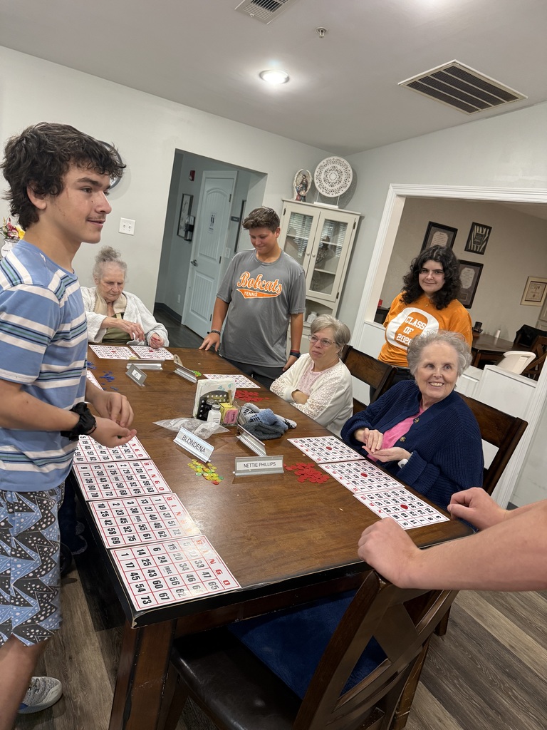 Several people, including kids, are seated around a table playing a game. One person stands and looks at the game board.