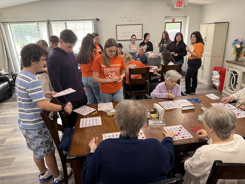 A group of people wearing orange shirts sit around a table with cards. Some sit on chairs, others stand, all facing the table.