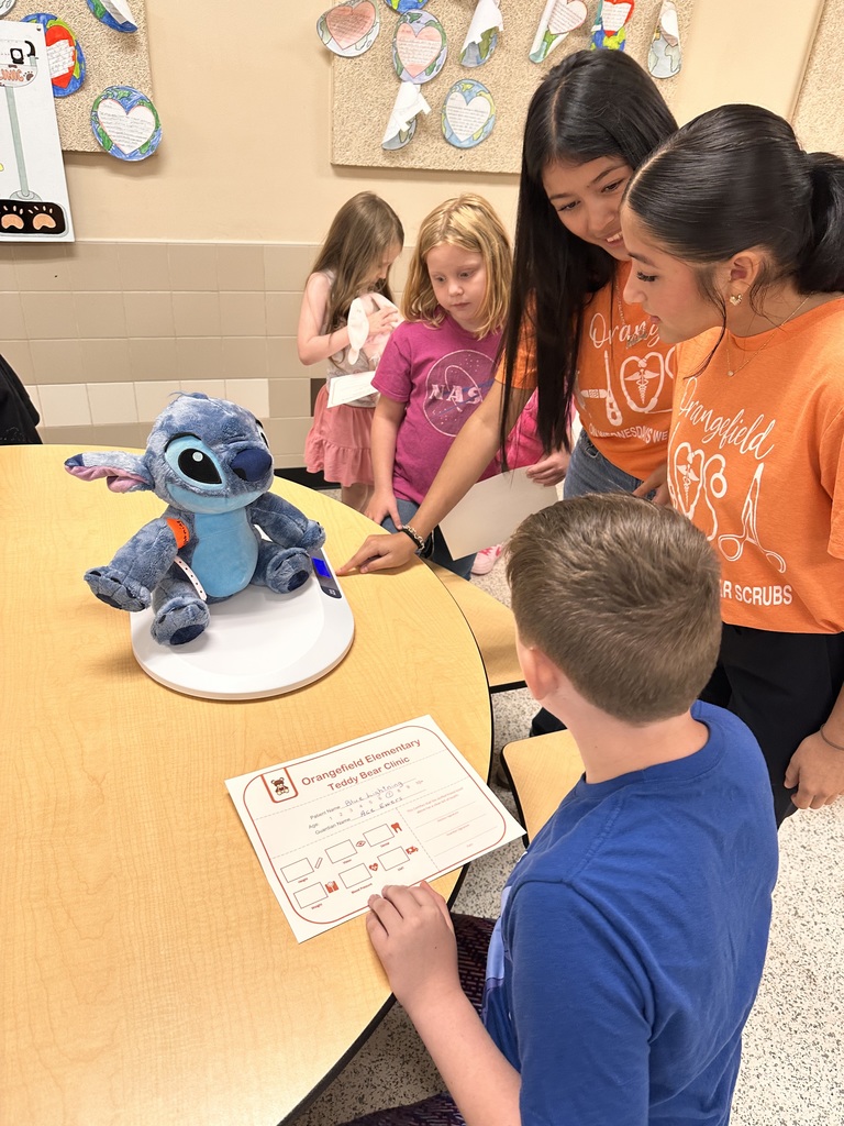 A stuffed animal is on a table. Two teenagers in orange shirts assist a kid with a worksheet.