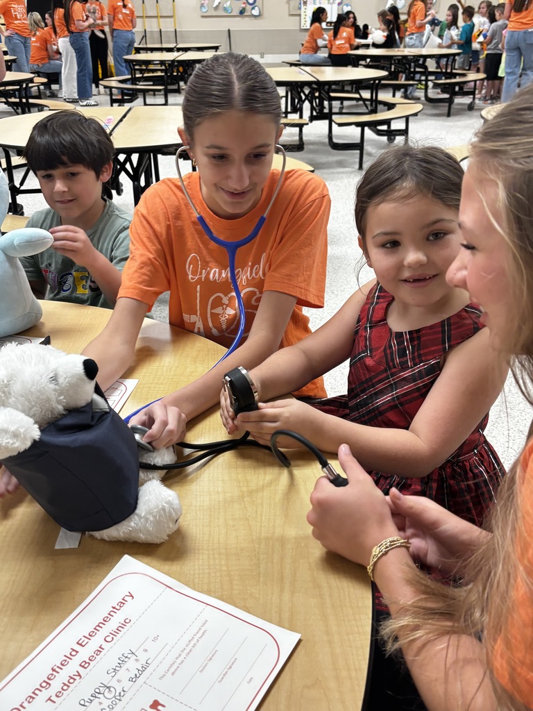 Kids using a stethoscope on a stuffed toy. One wears an orange shirt, another a red dress.