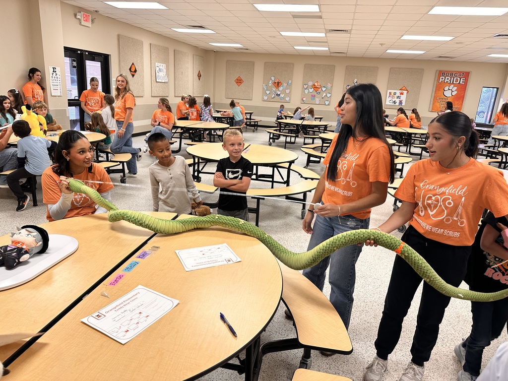 Several children in orange shirts interact with a large green snake on a table in a classroom.