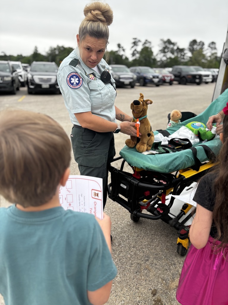 A woman in a uniform holds a stuffed toy as two children watch her. They are in a parking lot with vehicles.