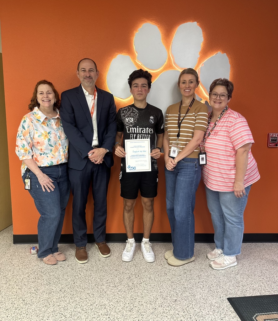 Five adults and a young man stand in front of an orange wall. The man holds a certificate.