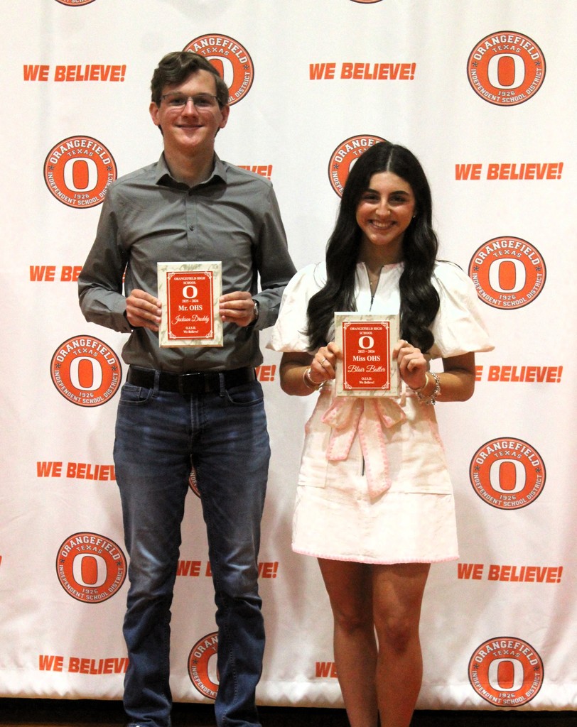 Two individuals stand with awards in a room with a white backdrop displaying the words "WE BELIEVE!" and circular logos.