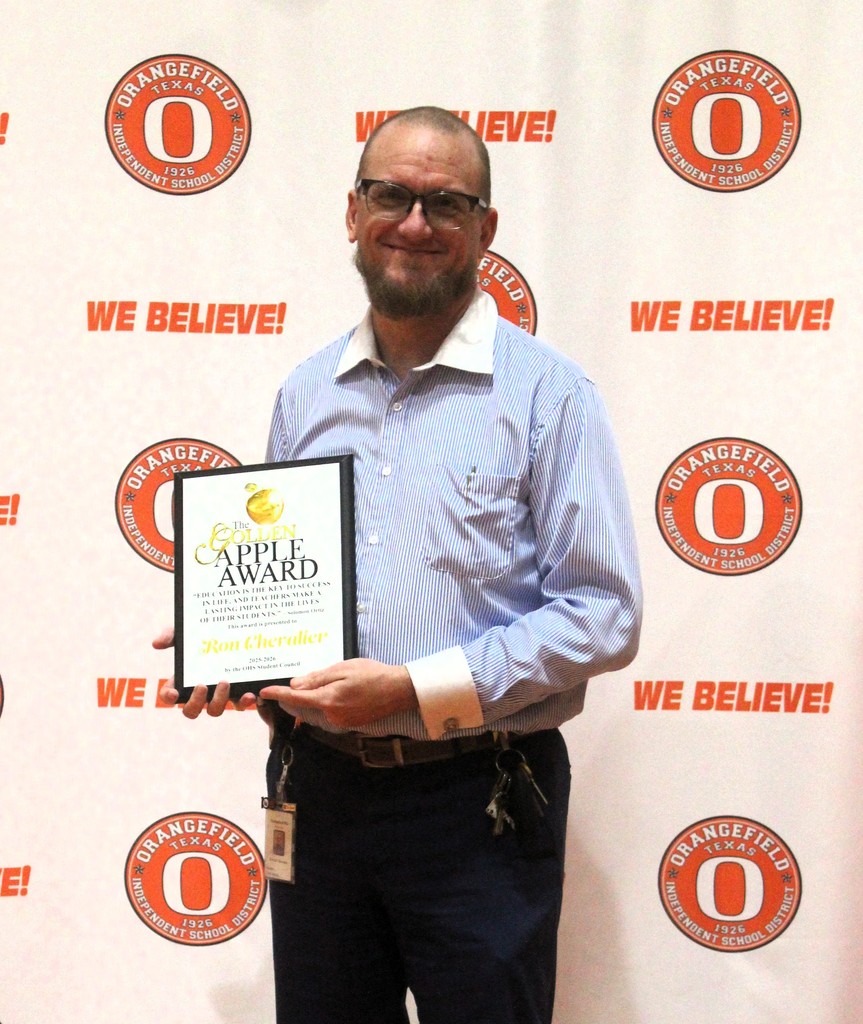 A man stands with a plaque in hand, smiling for a photo. Behind him, a backdrop displays "Orangefield We Believe!" in orange and white.
