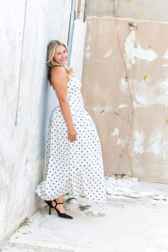 Woman in polka-dot dress, black heels, and a smile standing against a peeling wall. Charlee