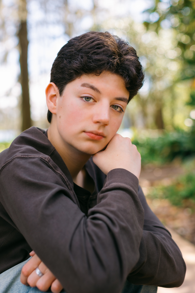 A boy with short hair sits outdoors with one arm resting on his knee and his hand under his chin. Landon