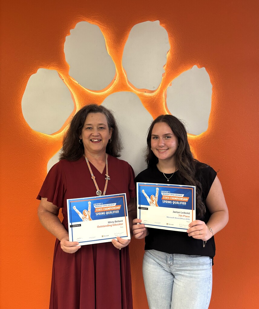 A teacher and student pose next to each other holding certificates. Behind them is an orange wall with a large paw print cutout.