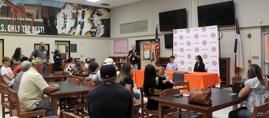 Group of people seated around tables in a room. A person stands near a banner. Orange tablecloths.