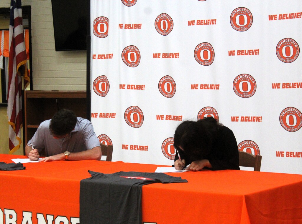 Two individuals writing on paper at a table with an orange tablecloth and a white backdrop with orange lettering.