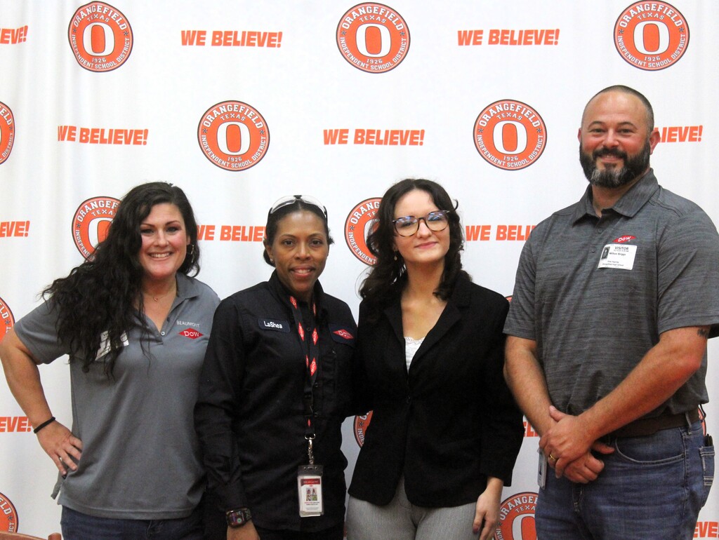 Four people stand together in front of a wall with large, round orange signs featuring the text "WE BELIEVE!" and a white "O" symbol.