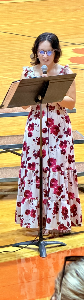 Student in a white dress with red flowers singing into a microphone  