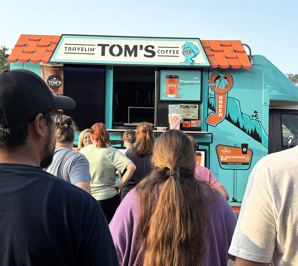 A crowd of people stand in front of a Tom's Coffee truck. The truck is blue and orange.