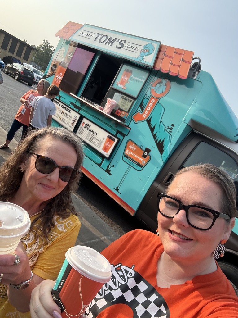 Two women in front of a blue coffee truck. One woman has a cup in hand, and the other holds a cup with a checkered designed shirt.