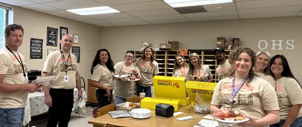 Group of people in matching shirts, some holding plates of food, gathered around a table with yellow boxes.