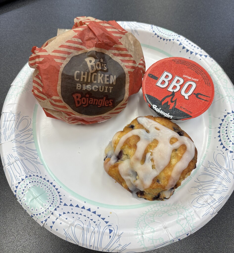 A white plate has a biscuit, a chicken sandwich, and a small container with a red lid.