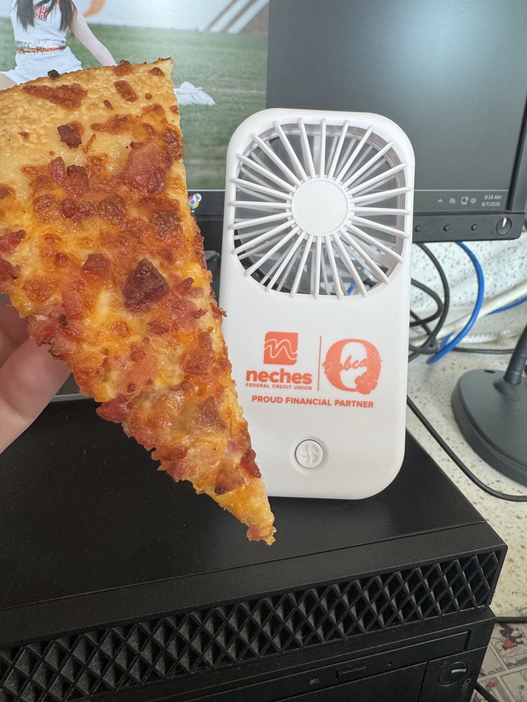 A slice of pizza is held in front of a small, white, fan-like device with branding on it. A computer monitor is in the background.