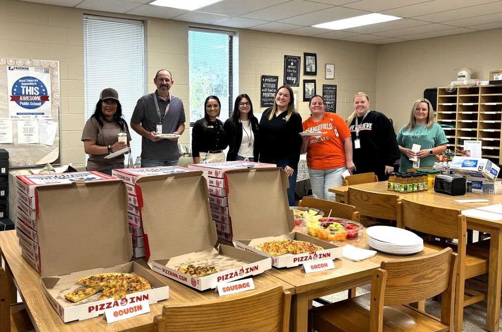 A group of people standing behind a table with pizza boxes, plates, and fruit in a workroom.