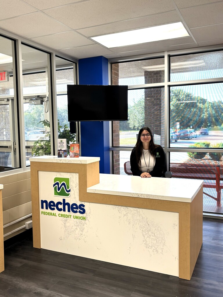 A person sits at a kiosk desk with a sign reading "Neches Federal Credit Union." The space has glass walls, a television, and a view of a parking lot.
