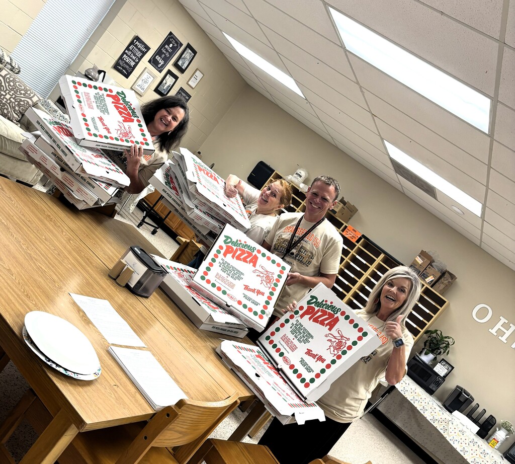 Four people in a room holding boxes of pizza, standing around a table. A sign reading "OHS" on the far right.