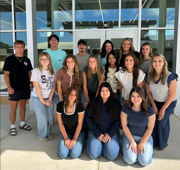 A group of students, both male and female, pose in front of a building with large glass windows.