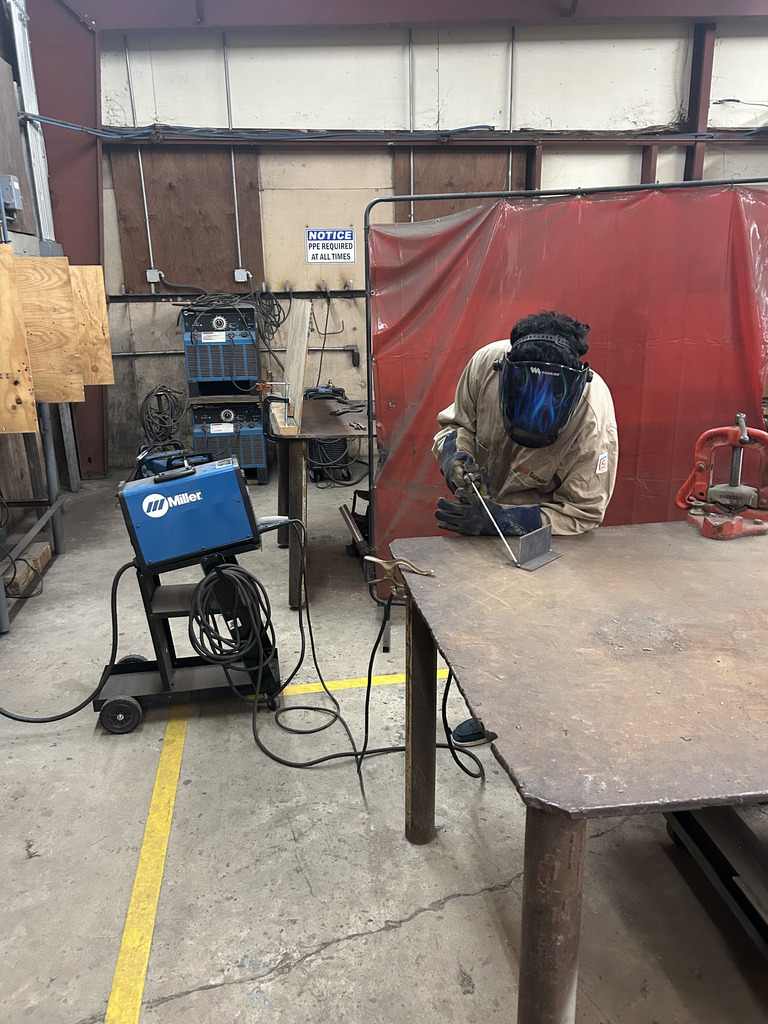 A welder in a welding shop, wearing protective gear, works on a metal surface using welding equipment.
