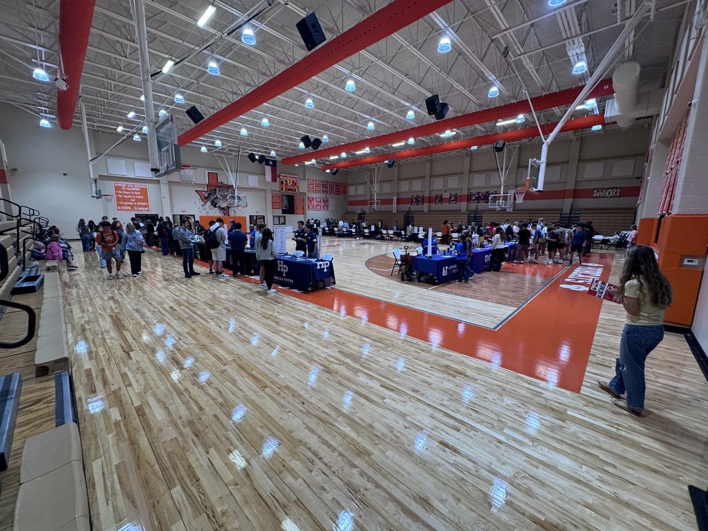 An indoor basketball court with wooden floors and bleachers. People walk around tables and a woman stands near the court.