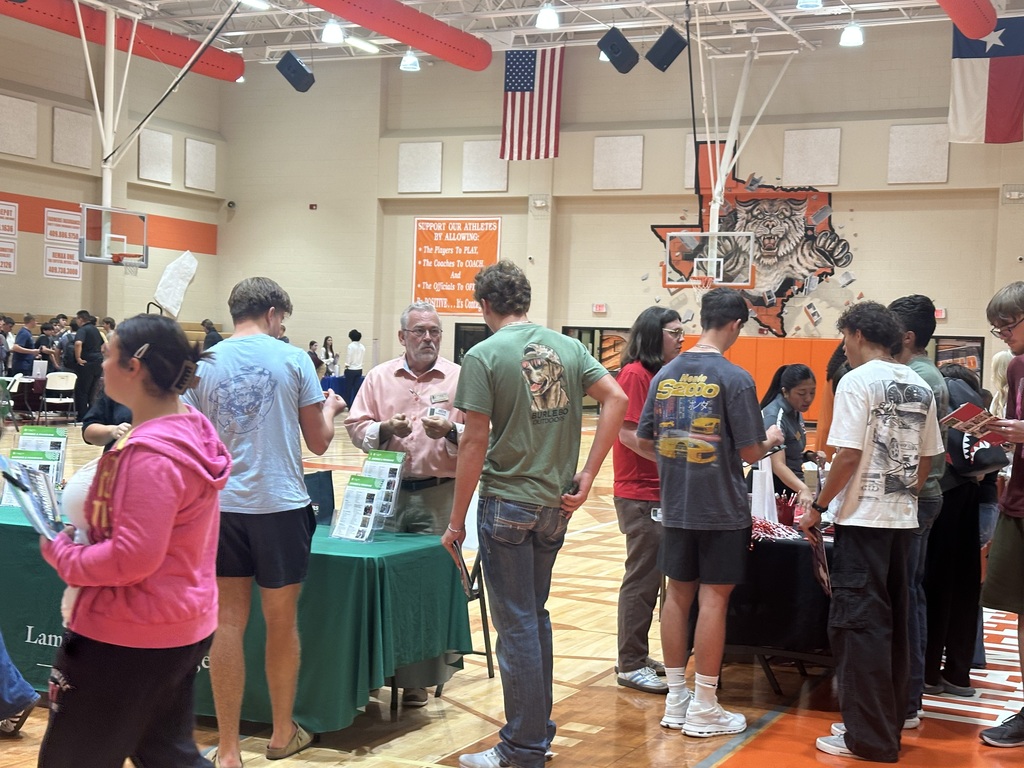 A group of people stand around tables in a gymnasium with banners on the walls and a basketball hoop in the background.