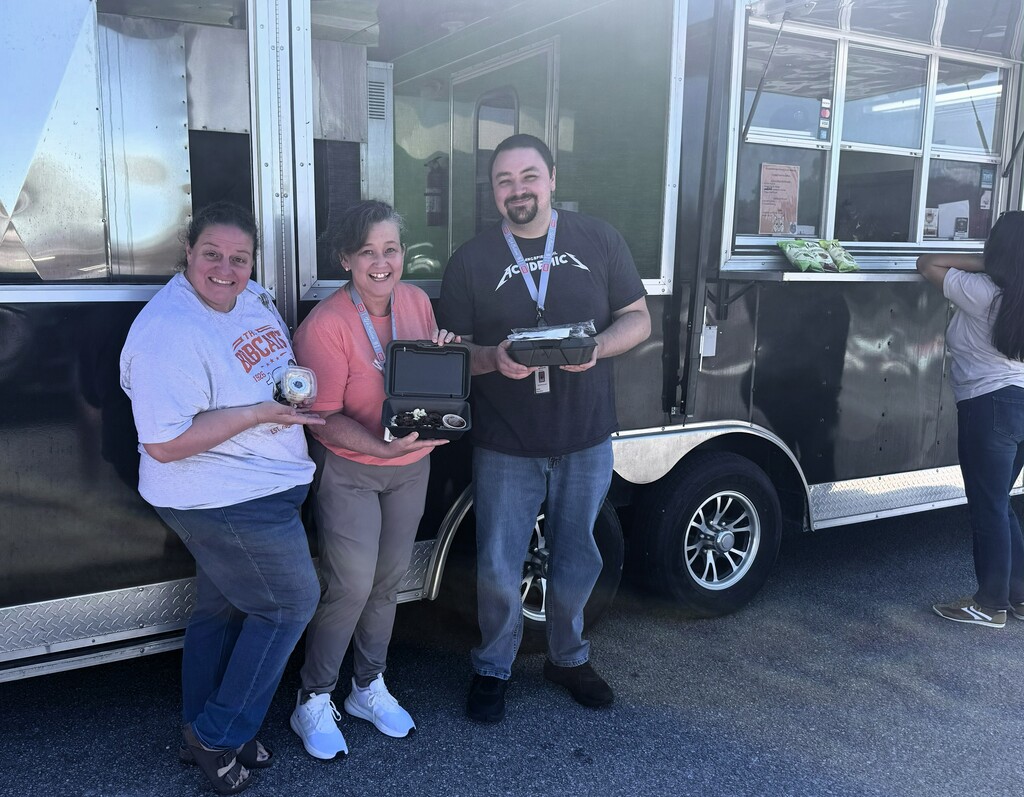 Three people pose with items in front of a parked food truck. The man wears a lanyard.