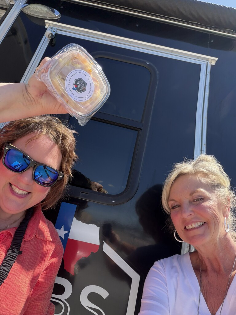 Two women smiling, one holding a clear container with food, standing by a black vehicle with a Texas flag design.