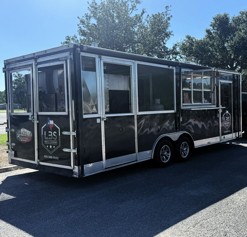 A black food truck with glass windows parked on the side of the road. Trees and grass in background.