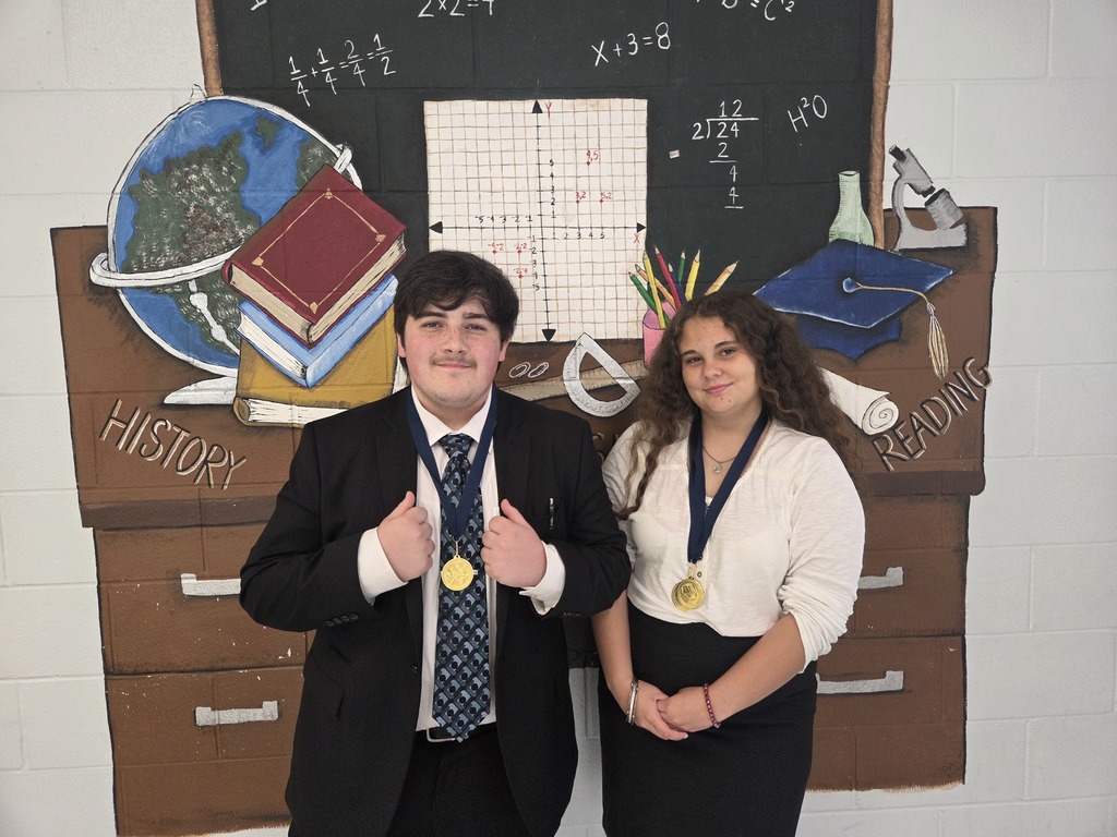 A man and a woman, both wearing medals, stand in front of a decorated classroom backdrop.