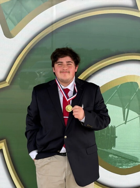 A student in a black suit and red tie stands in front of a green and gold sign, holding a gold medal.