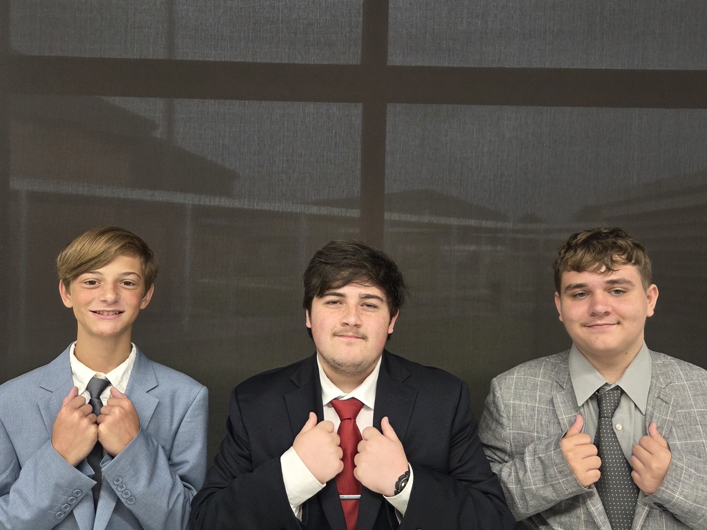 Three young men dressed in formal suits, standing close together. They are adjusting their ties.