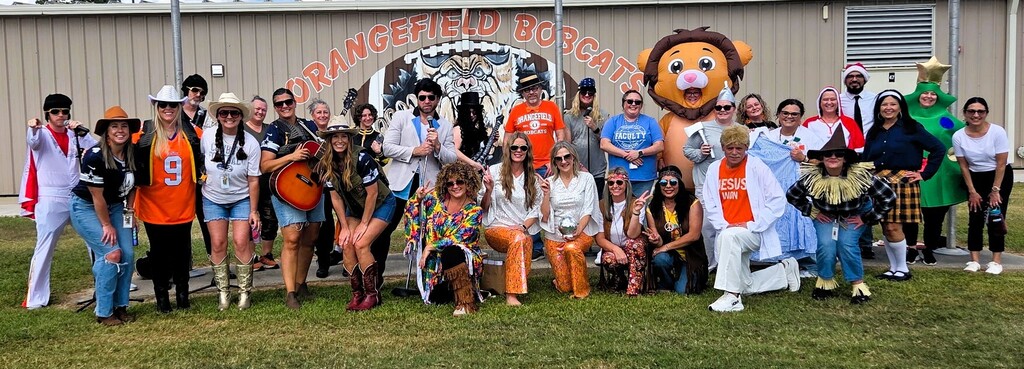 A group in costumes stands in front of a building. Some wear hats and boots; others wear costumes.