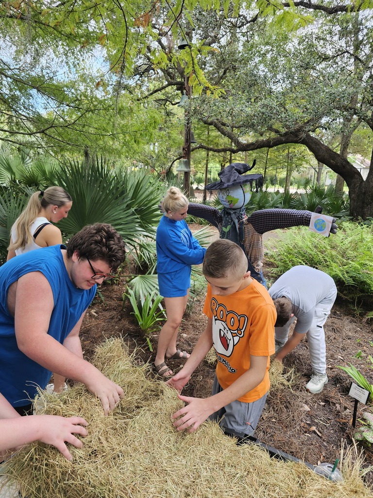 A group of people gather around a haystack, possibly participating in a community project. They work together in a garden with trees and plants.