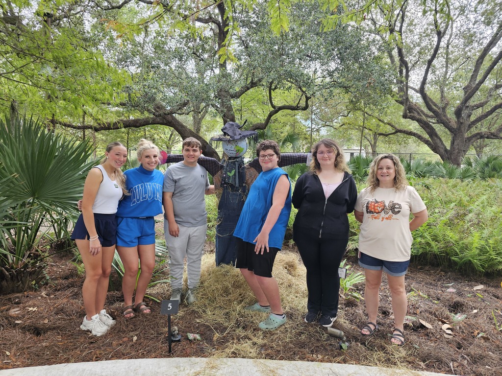 Six people stand in a garden with greenery and trees in the background.