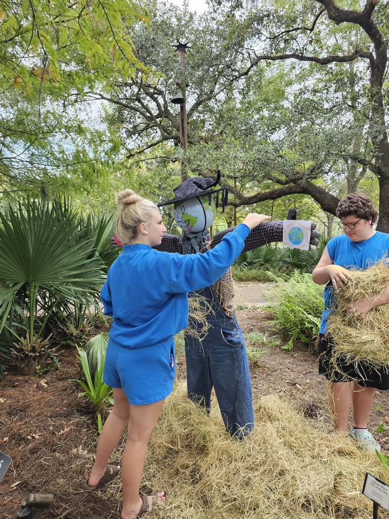 Three people in a garden, one dressing a scarecrow, another holding hay, with green trees and plants.