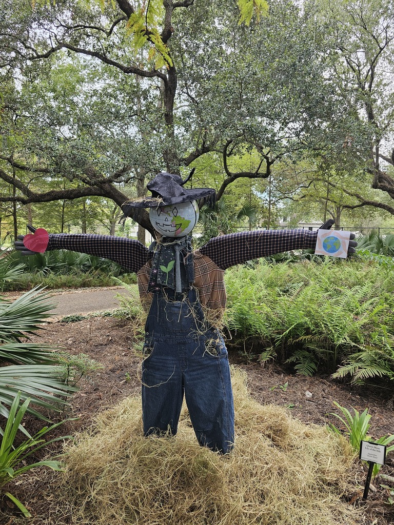 A scarecrow wearing a hat, overalls, and a mask stands in a garden with green foliage.