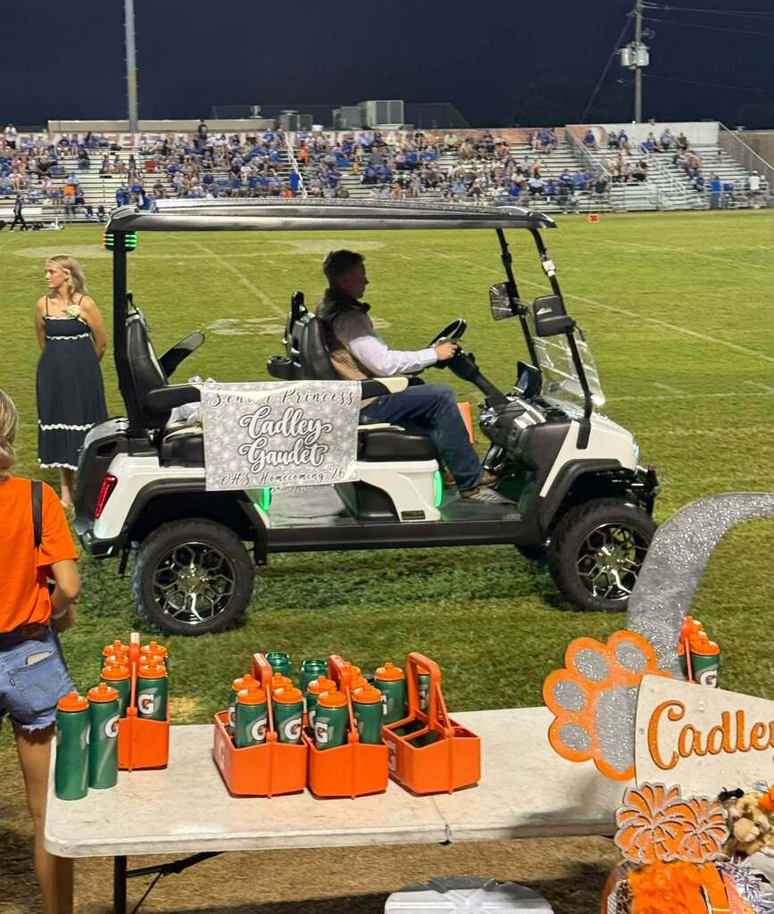 Golf cart parked on a football field with refreshments on a table. A woman stands near the cart.